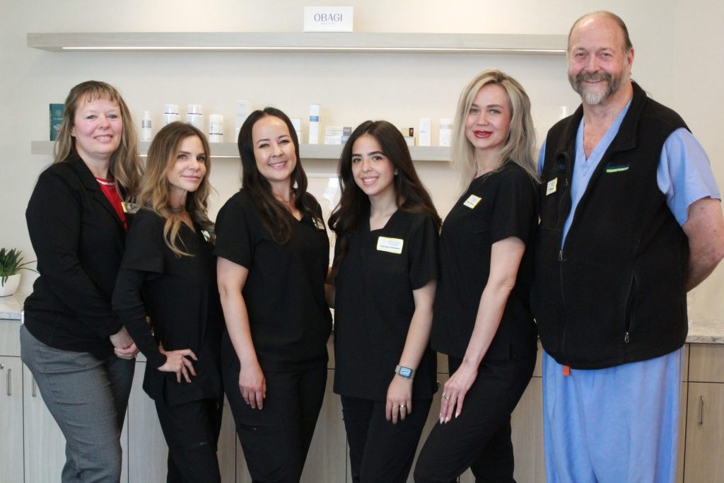 Six smiling people stand in a row in a clinic, five women in black uniforms and one man in blue scrubs and a black vest. Shelves with skincare products promoting anti-aging and wellness are on the wall behind them, highlighting the commitment of the Institute For Ageless Living.