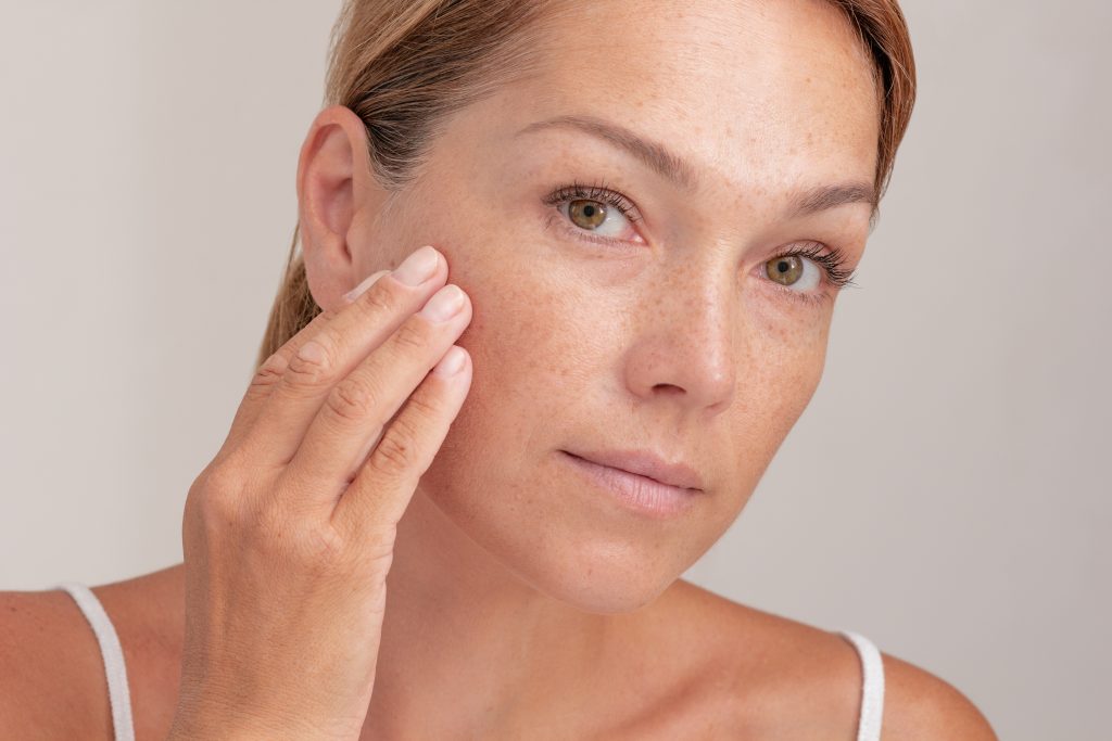 A woman with light skin and freckles touches her cheek gently, looking directly at the camera with a neutral expression. She has light brown hair pulled back and wears a white tank top, embodying the natural confidence promoted by the Institute For Ageless Living's approach to wellness.
