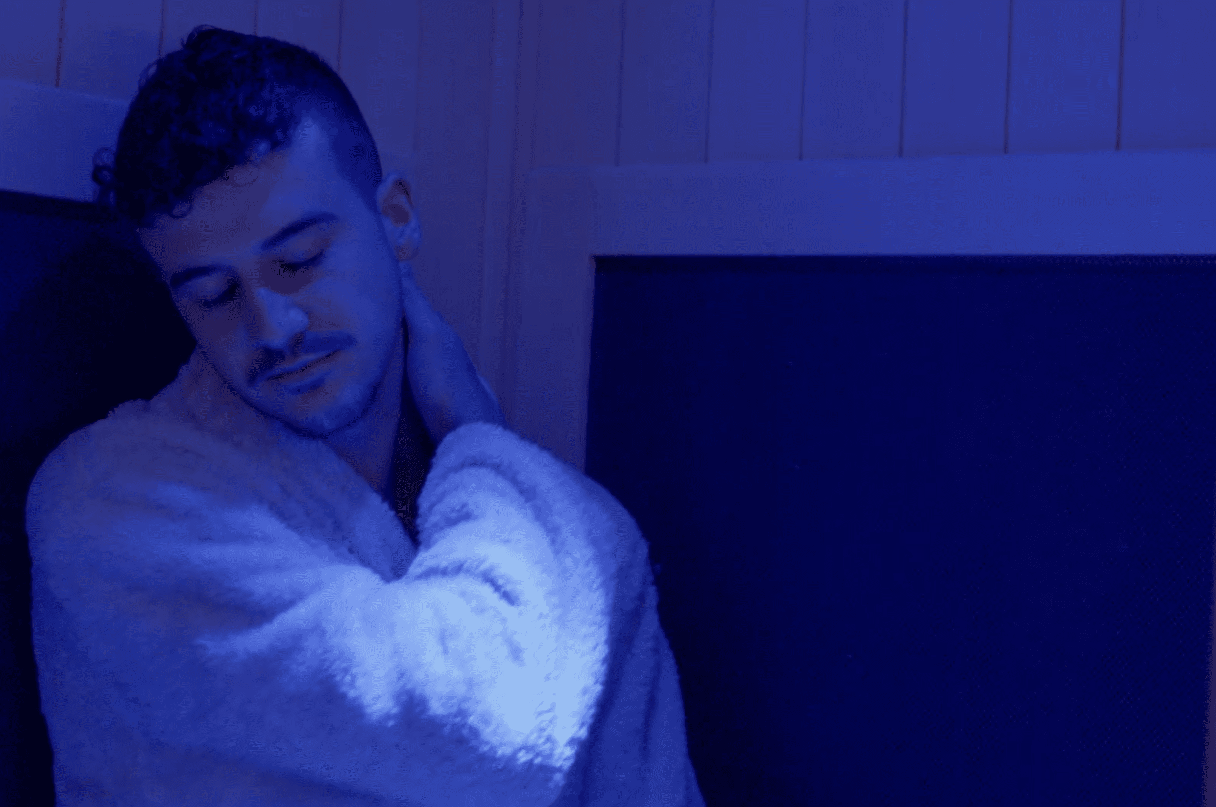 A person wearing a robe stands in blue lighting, eyes closed and head tilted, gently touching the back of their neck with one hand—capturing a serene moment inspired by the wellness philosophy of the Institute For Ageless Living.