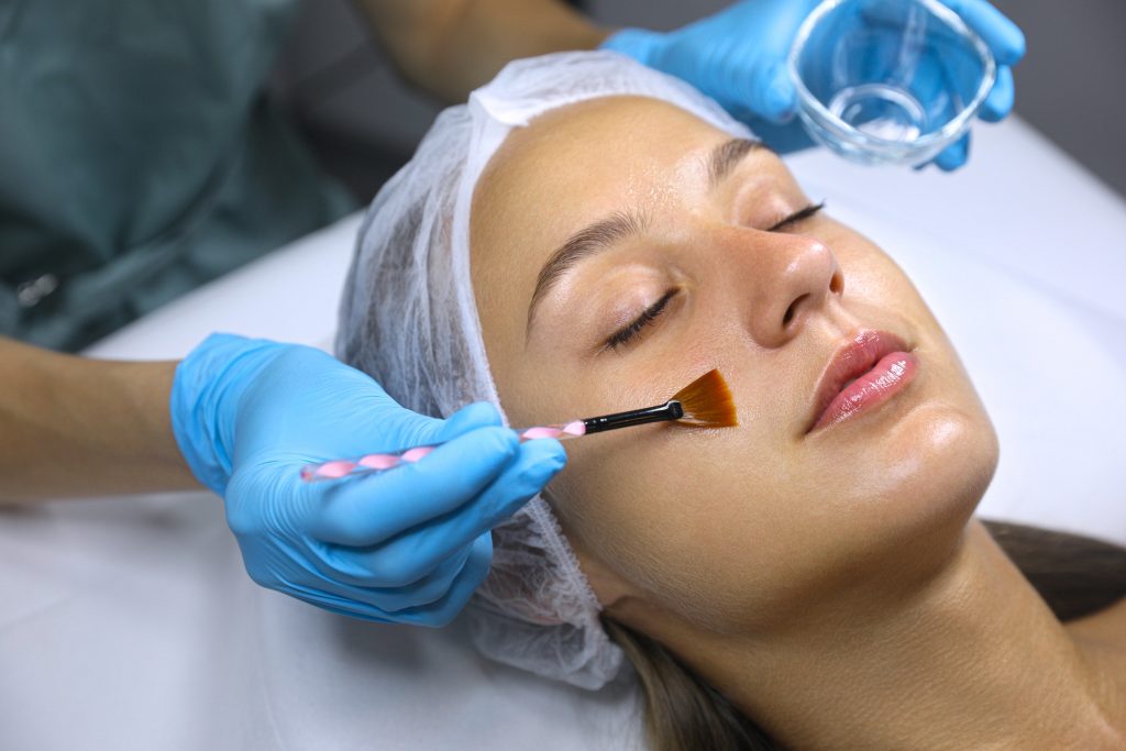 A person with a hair cover receives an anti-aging facial treatment as someone wearing blue gloves applies a clear liquid to their cheek with a brush, promoting wellness in line with the standards of the Institute For Ageless Living.