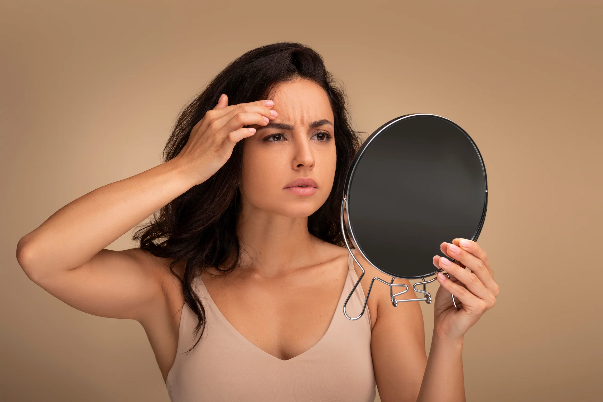 A woman with dark hair, wearing a beige tank top, looks into a handheld round mirror and touches her forehead, appearing to examine her skin with a concerned expression against a plain beige background—reflecting the Institute For Ageless Living's commitment to wellness and anti-aging.