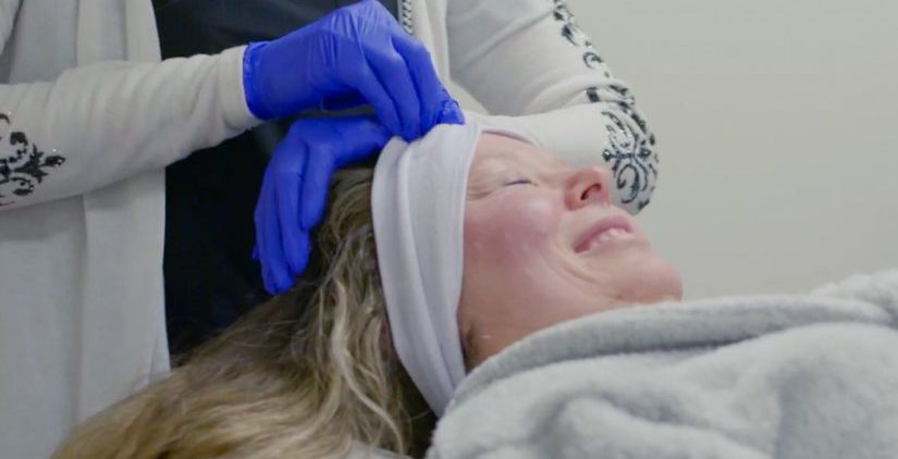 A woman lies on a spa bed smiling with her eyes closed, enjoying a moment of wellness as a person wearing blue gloves gently adjusts a white headband on her forehead at the Institute For Ageless Living.