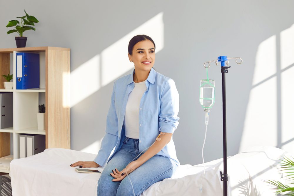 A woman sits on a medical bed at the Institute For Ageless Living, smiling, with an IV drip attached to her arm. She wears a light blue shirt and jeans. Sunlight streams through a window, and there are plants and shelves in the background, creating an inviting wellness environment.