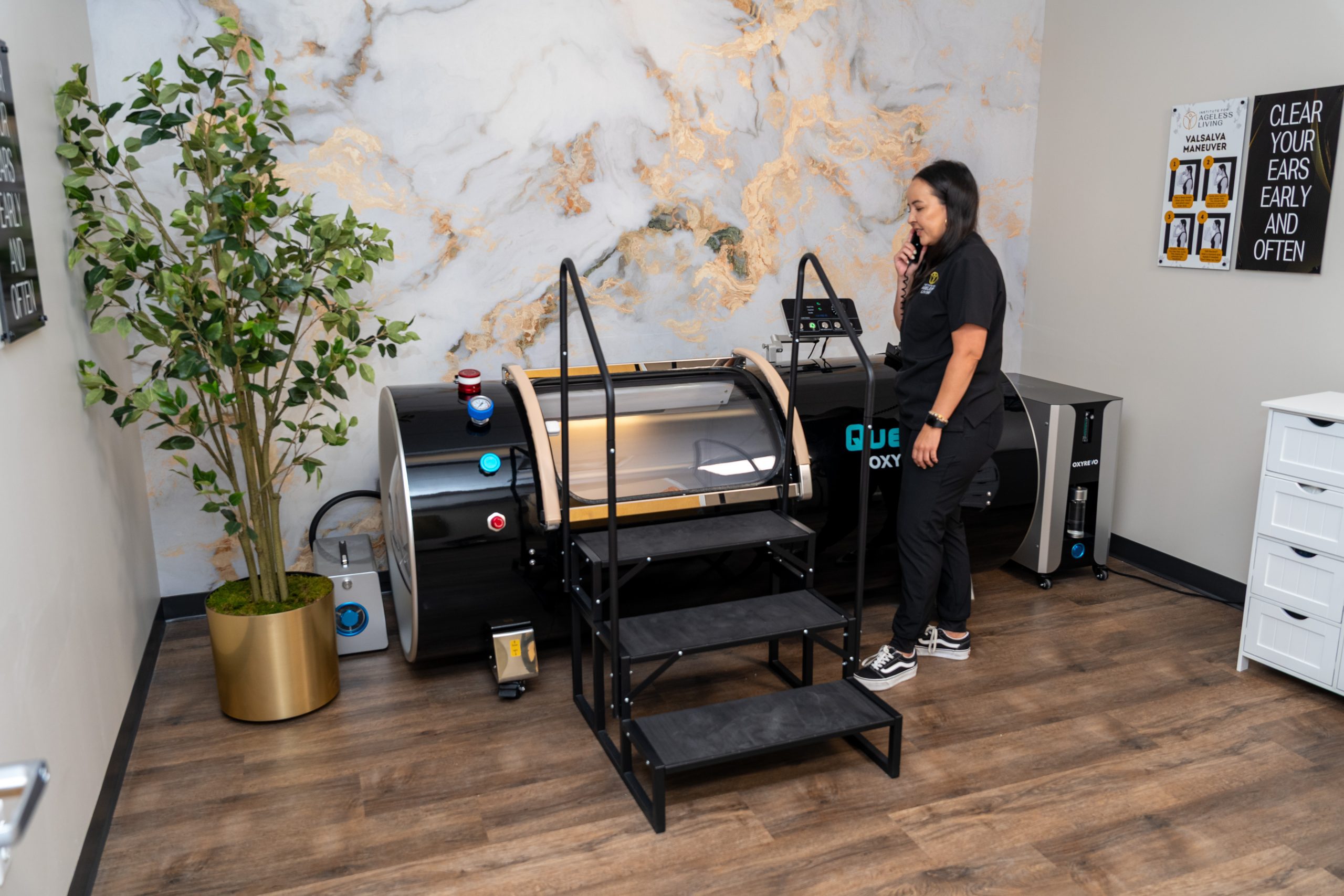 A person stands beside a large hyperbaric oxygen chamber in a modern room with wood floors, a potted plant, steps leading to the chamber, and a marble-patterned wall—an inviting space dedicated to wellness and anti-aging at the Institute For Ageless Living.