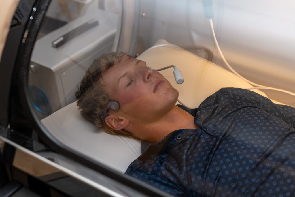 A young man lies inside a transparent chamber, eyes closed, wearing a patterned shirt and small headsets on his head. The scene appears clinical or medical, suggesting an advanced anti-aging procedure or wellness therapy session at the Institute For Ageless Living.