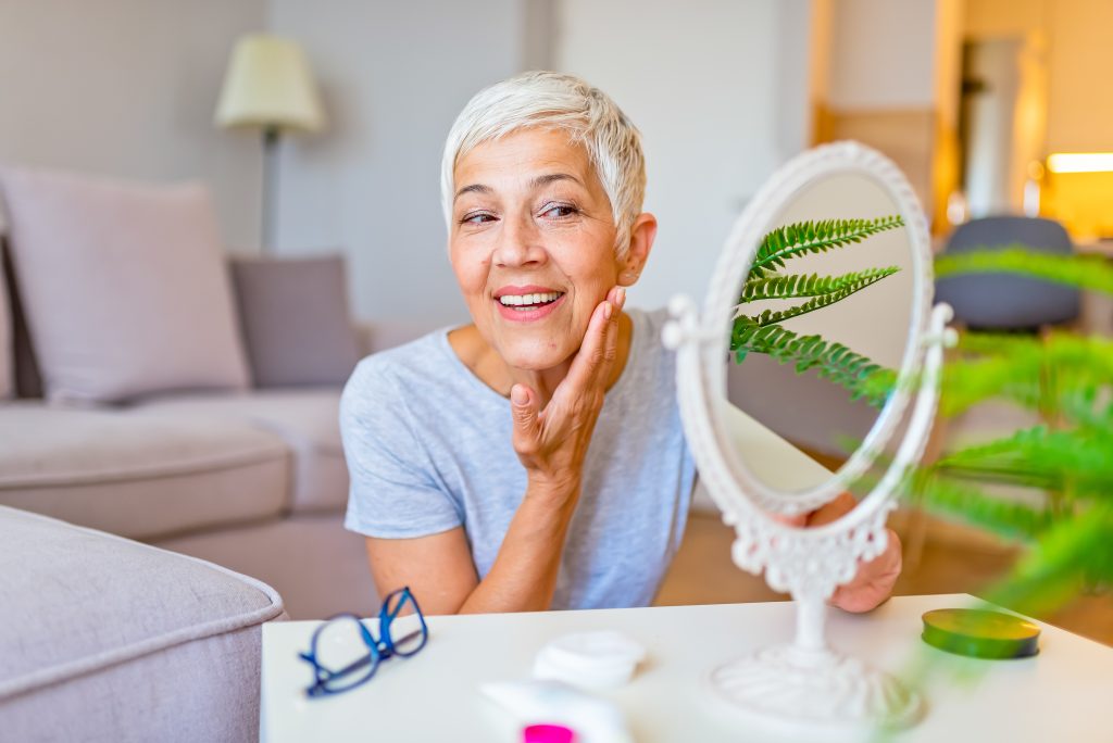 Smiling older woman with short gray hair touches her face while looking in a round mirror at home. Glasses and skincare items from the Institute For Ageless Living are on the table, highlighting her dedication to wellness and anti-aging, with green leaves in the foreground.