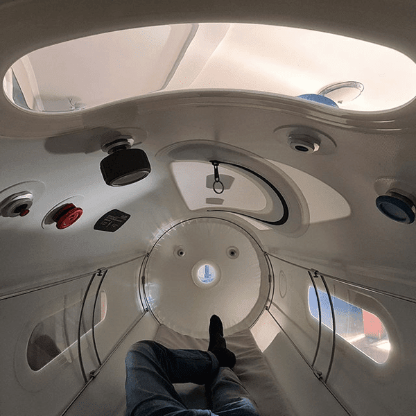 A person lies inside a white hyperbaric oxygen chamber, with various controls and windows visible overhead. The cylindrical chamber is designed to promote wellness, and the person's legs and one foot are in view. This setting is reminiscent of treatments offered by the Institute For Ageless Living, where advanced therapies support vitality and anti-aging benefits.