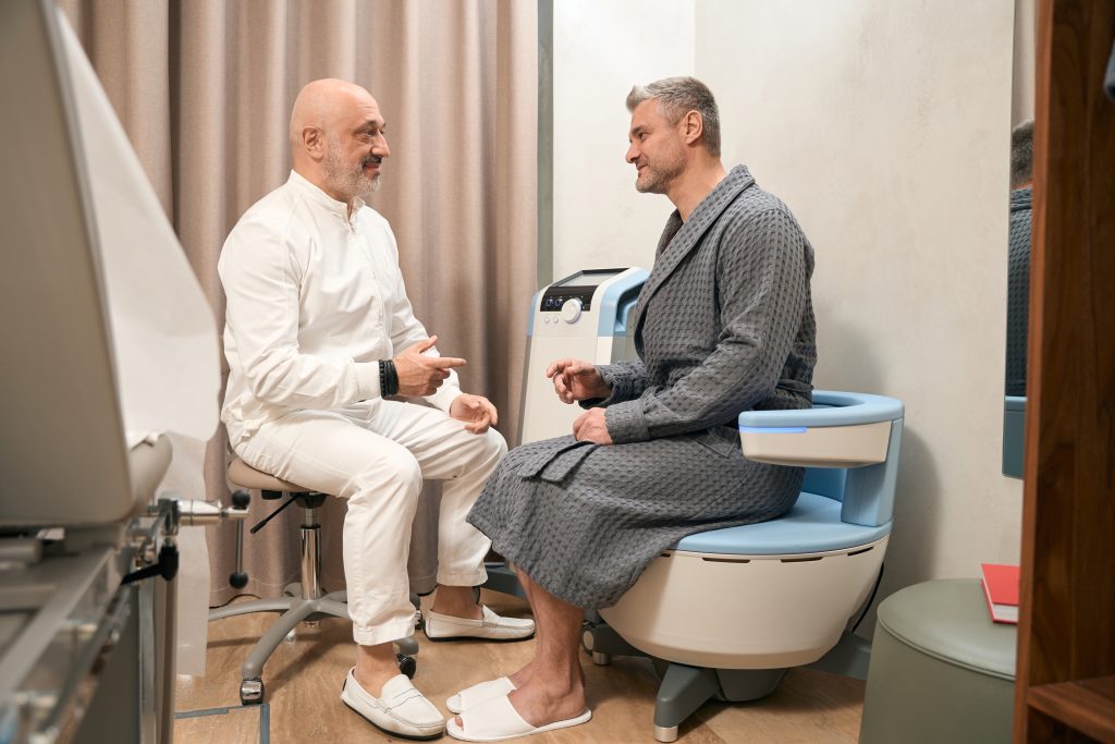 A doctor in white talks with a patient in a blue robe sitting on a medical chair. They appear to be having a wellness consultation in a modern clinic room at the Institute For Ageless Living.