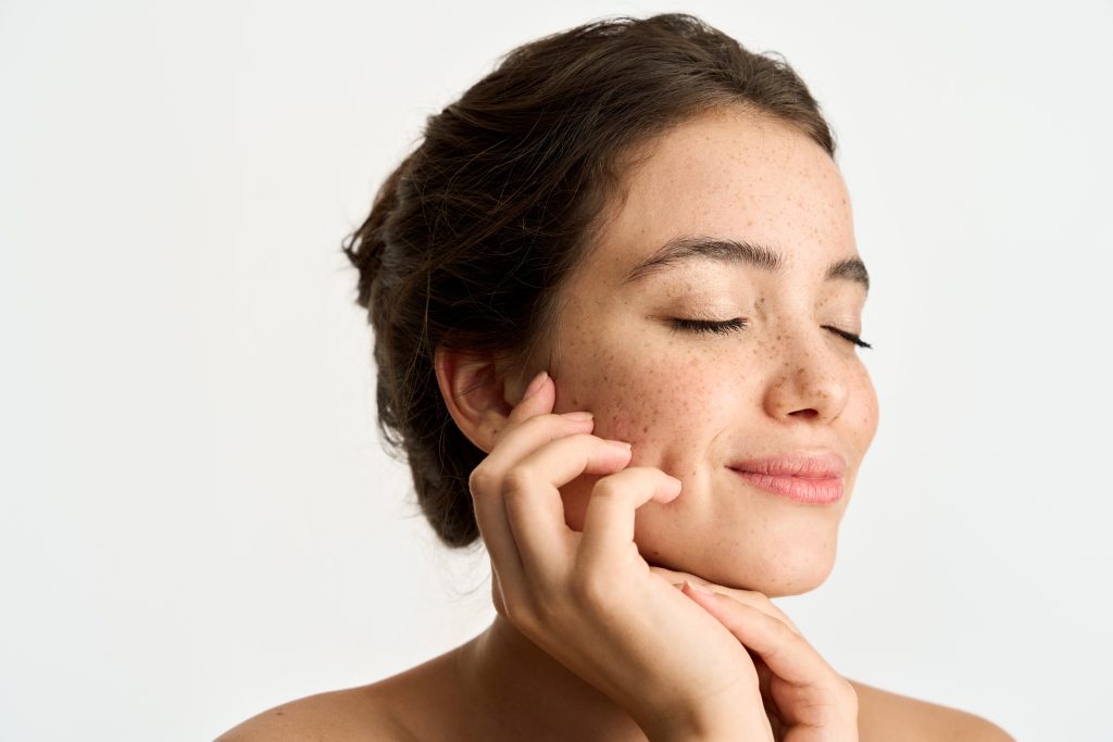 A woman with brown hair and freckles smiles gently with her eyes closed, lightly touching her face with both hands against a plain white background, embodying the spirit of wellness promoted by the Institute For Ageless Living.