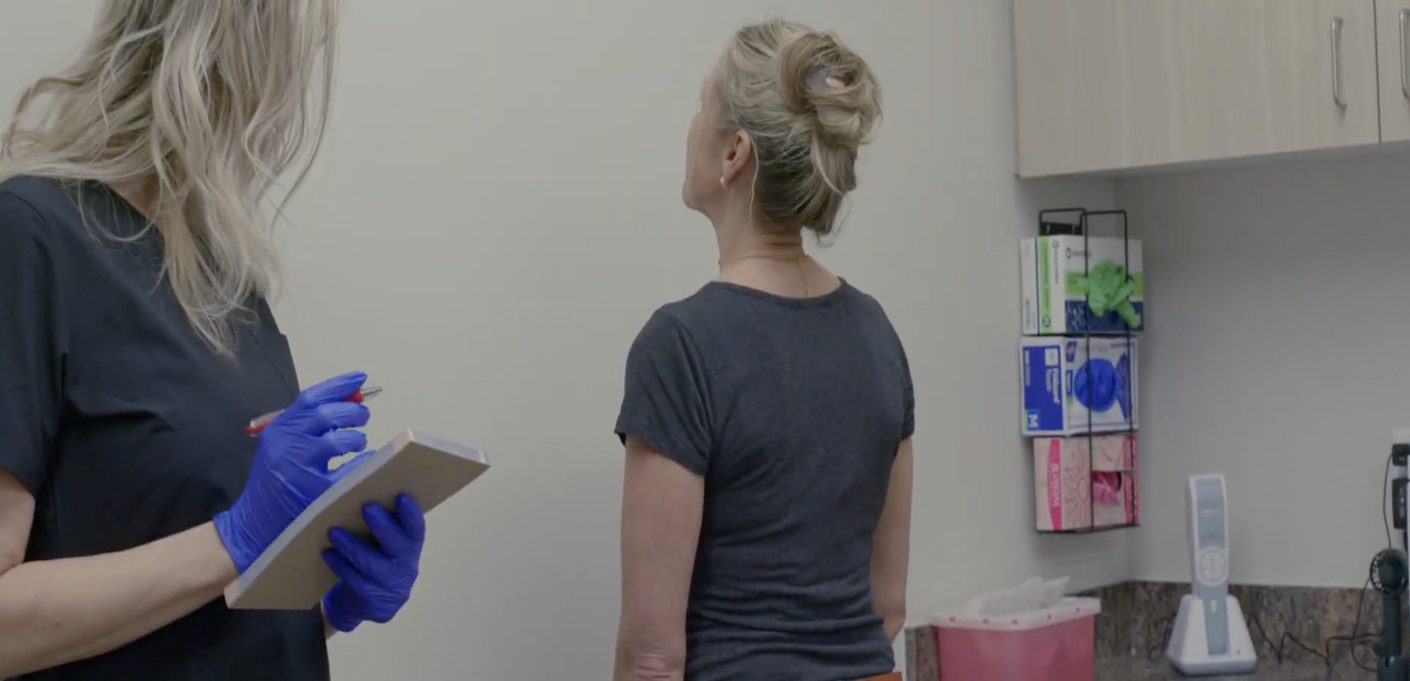A woman with her hair in a bun stands facing a wall in a medical office at the Institute For Ageless Living, while a healthcare worker in blue gloves writes on a clipboard. Medical supplies and a phone are visible on the counter, reflecting the clinic's commitment to wellness and anti-aging care.
