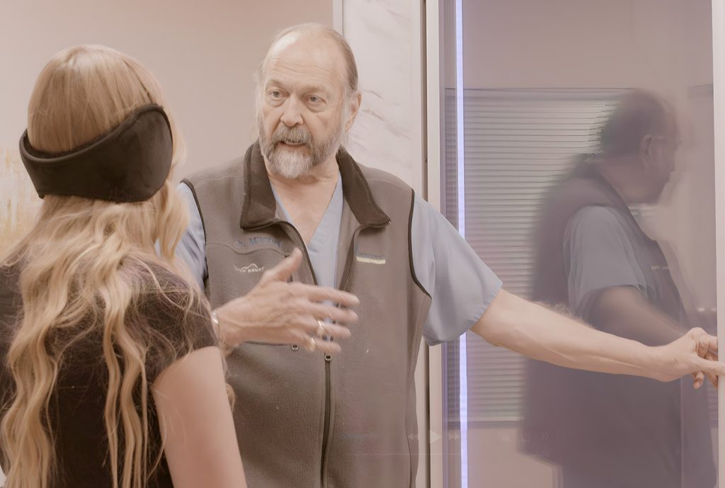 A man in medical scrubs and vest gestures while talking to a woman with long blonde hair and black earmuffs in a clinic setting, discussing wellness strategies at the Institute For Ageless Living.