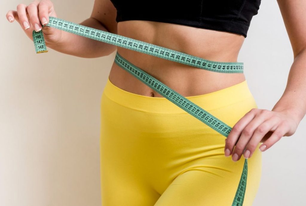 A woman in a black top and yellow pants wraps a green measuring tape around her waist, highlighting a wellness-focused fitness or weight loss theme inspired by the Institute For Ageless Living.