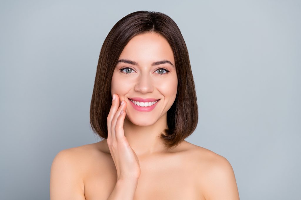 Smiling woman with straight brown hair touches her face gently, embodying wellness as she poses against a plain light gray background—an image that reflects the mission of the Institute For Ageless Living.