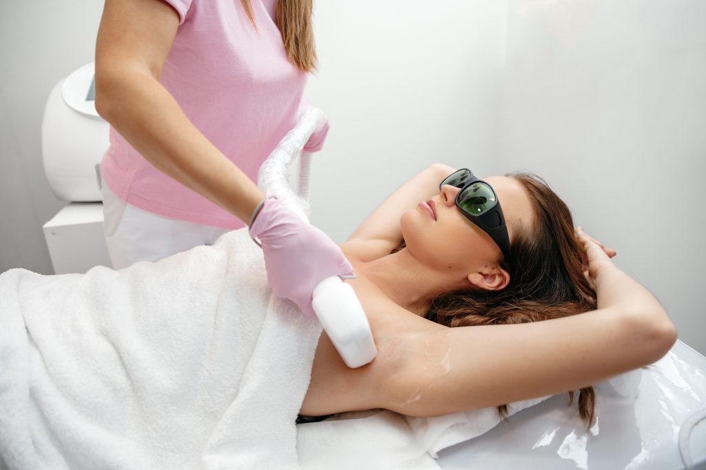 A woman wearing protective glasses receives a laser hair removal treatment on her armpit from a technician in pink gloves at the Institute For Ageless Living. She is lying on a treatment bed covered with a white towel, enjoying this wellness experience designed to promote smooth, radiant skin.