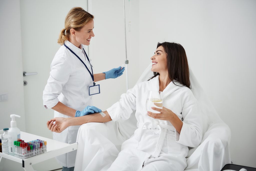 A healthcare professional prepares to draw blood from a woman in a robe, who is sitting in a chair and smiling while holding a glass of water. The scene takes place in the Institute For Ageless Living, reflecting a clean, bright medical setting dedicated to wellness and anti-aging care.