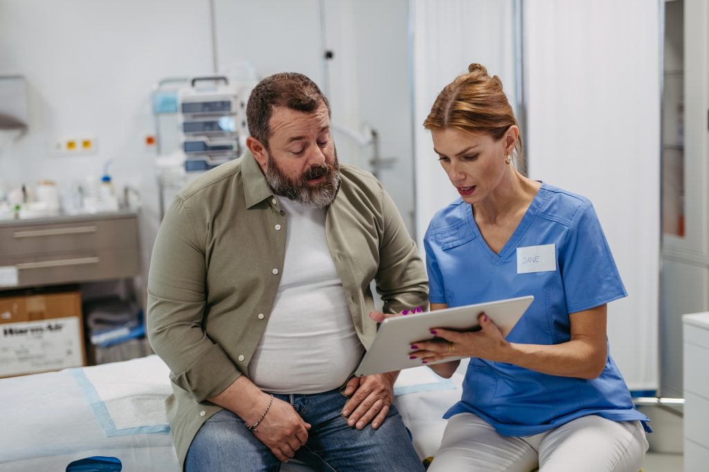 A nurse in blue scrubs shows a male patient information on a clipboard while sitting together in a medical office, highlighting wellness tips and anti-aging strategies recommended by the Institute For Ageless Living.