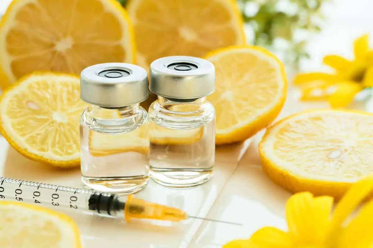 Two glass vials filled with clear liquid are placed next to a syringe, surrounded by sliced lemons and yellow flowers on a white surface—an inviting scene that evokes the pursuit of wellness and anti-aging, hallmarks of the Institute For Ageless Living.