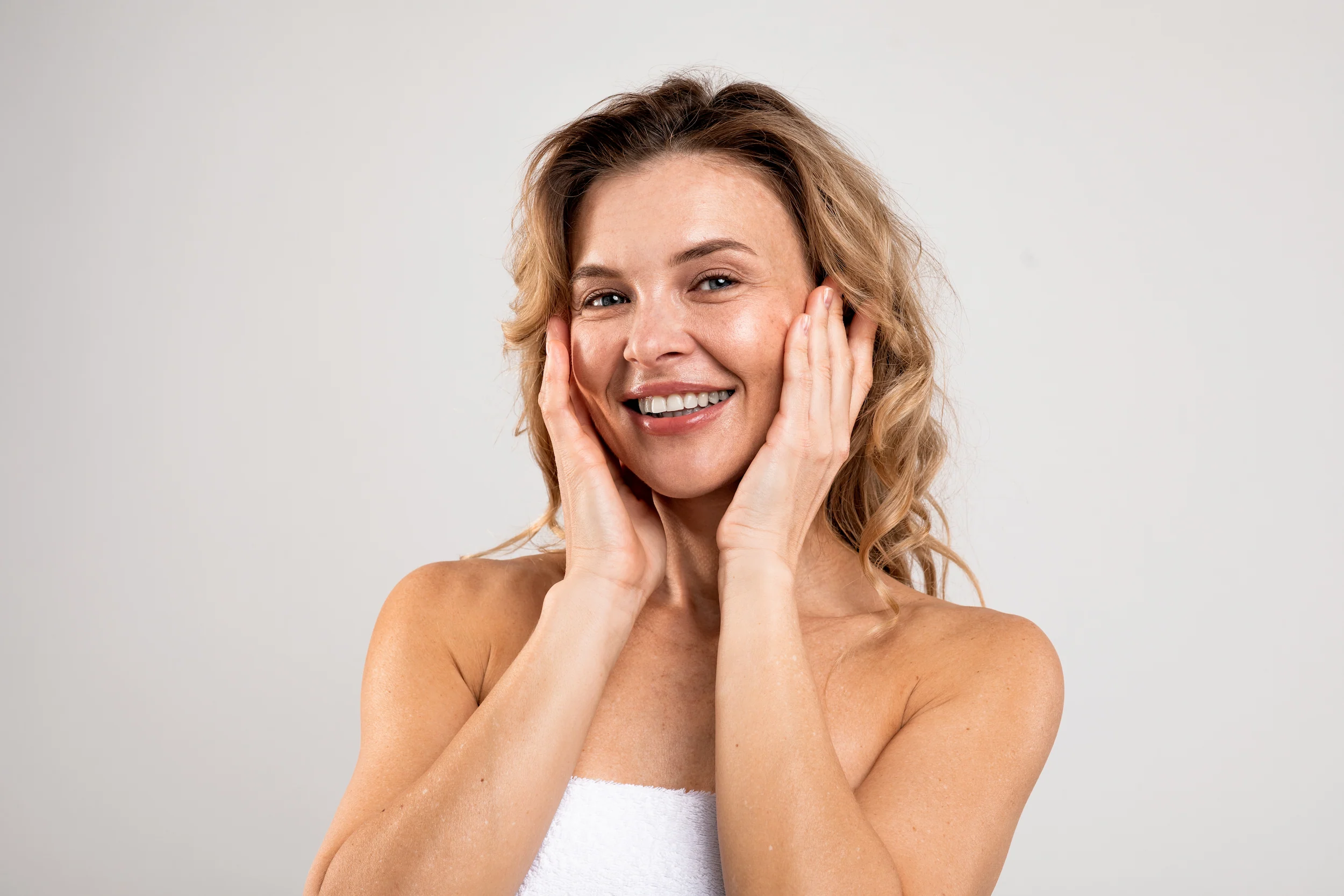 Smiling woman with wavy blonde hair holds her face with both hands, radiating a sense of wellness. She is wearing a white towel and stands against a plain light background, embodying the essence of anti-aging as promoted by the Institute For Ageless Living.