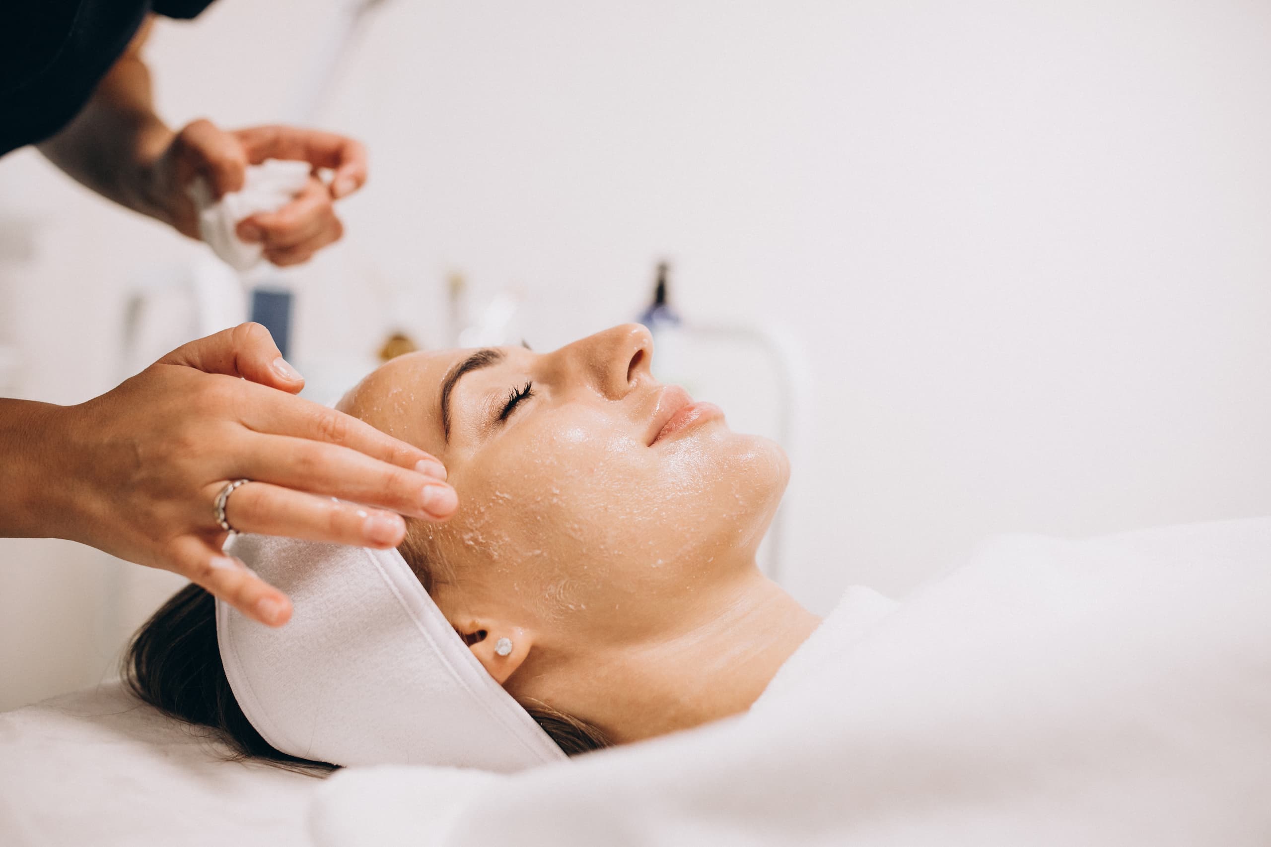 A woman lies on a spa bed with her eyes closed, receiving a luxurious facial treatment at the Institute For Ageless Living. An esthetician applies anti-aging cream to her face, promoting wellness and rejuvenation. The woman wears a white headband and is covered with a white towel. The setting is bright and clean.