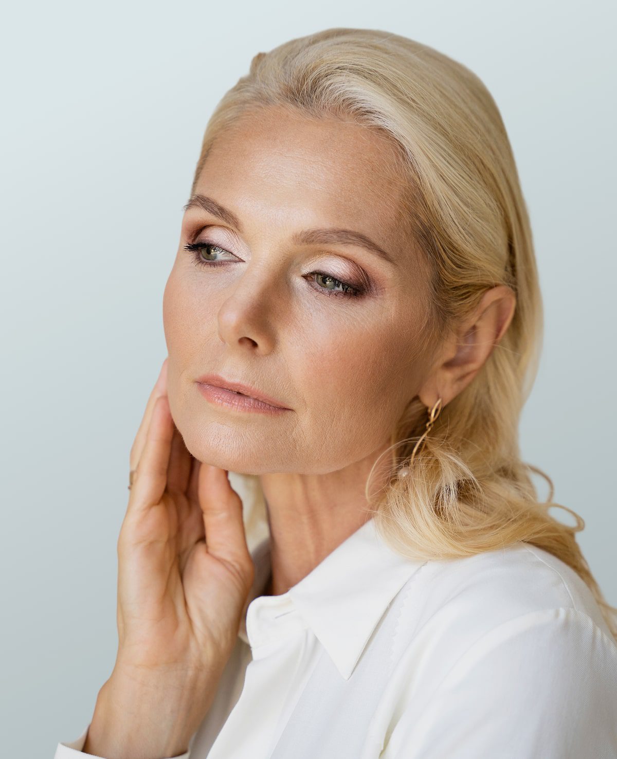 A woman with blonde hair and light makeup gazes thoughtfully off-camera, wearing a white collared shirt and gold hoop earrings, with one hand resting on her neck against a pale background—a serene portrait reflecting the essence of wellness promoted by the Institute For Ageless Living.