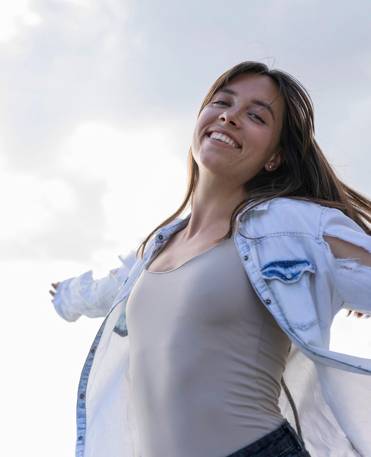 A young woman with long brown hair smiles joyfully with her arms outstretched, embodying wellness in a ripped denim jacket over a light tank top, standing outside against a bright, cloudy sky.