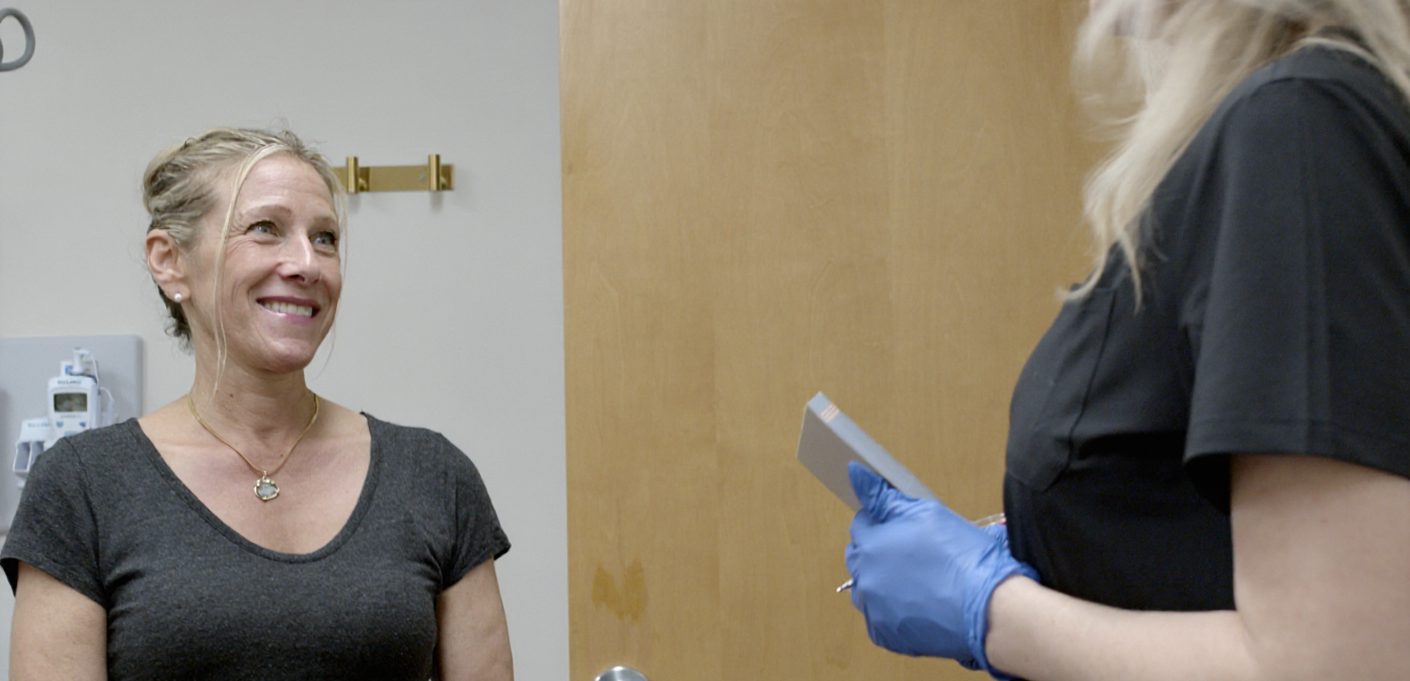 A woman in a gray shirt smiles at a healthcare worker wearing black scrubs and blue gloves, who holds a tablet. They appear to be in a medical office with a wooden door in the background, reflecting the welcoming environment of the Institute For Ageless Living dedicated to wellness and anti-aging care.