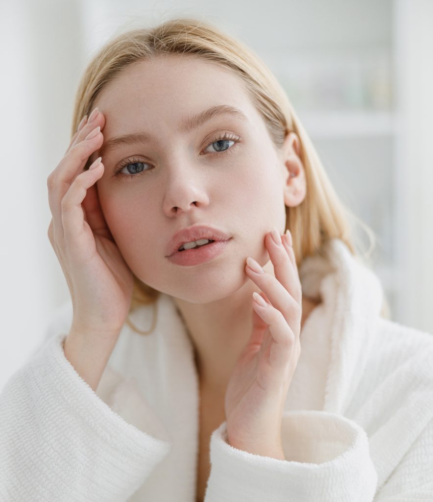A woman in a white bathrobe gently touches her face with both hands, looking directly at the camera with a calm, thoughtful expression. The background is softly blurred, suggesting a bathroom or spa setting—an atmosphere that perfectly embodies wellness and the anti-aging philosophy of the Institute For Ageless Living.