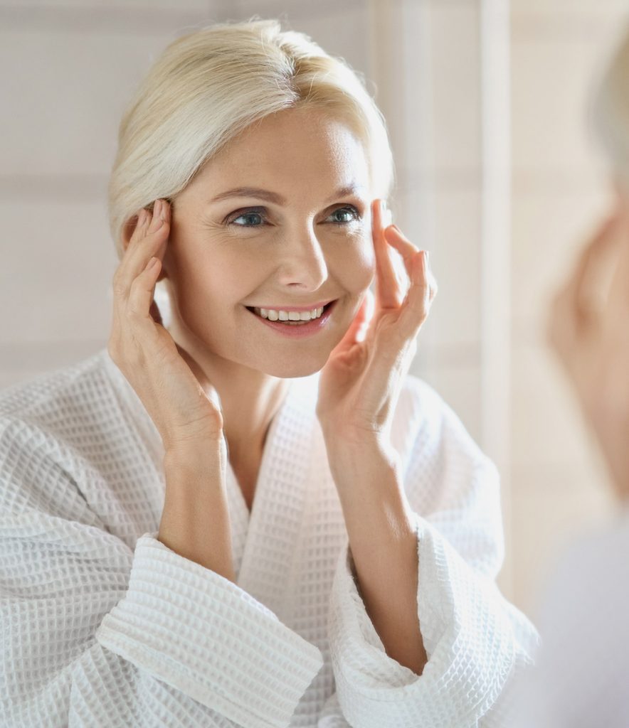A woman with light hair in a white robe smiles while touching her face and looking in the mirror, appearing happy and relaxed—radiating the wellness-inspired confidence promoted by the Institute For Ageless Living.