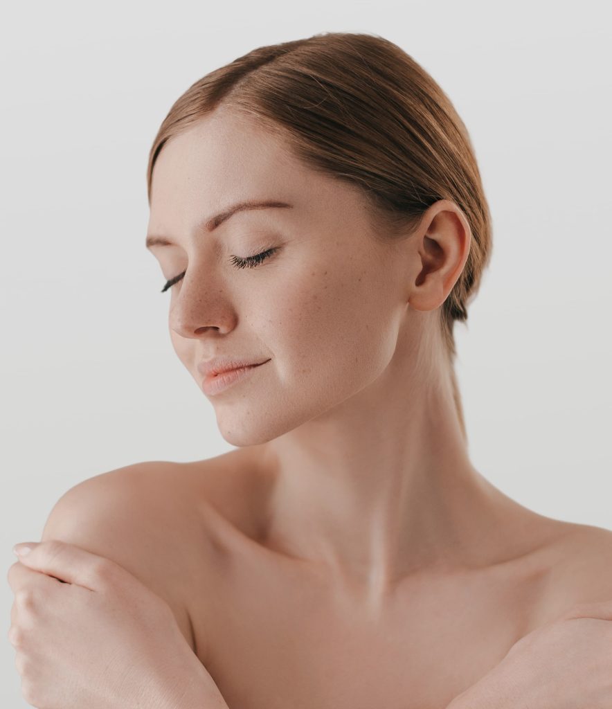 A young woman with fair skin and brown hair poses with eyes closed, gently touching her shoulder. The background is plain and light, creating a soft, serene atmosphere that embodies the essence of wellness and anti-aging.