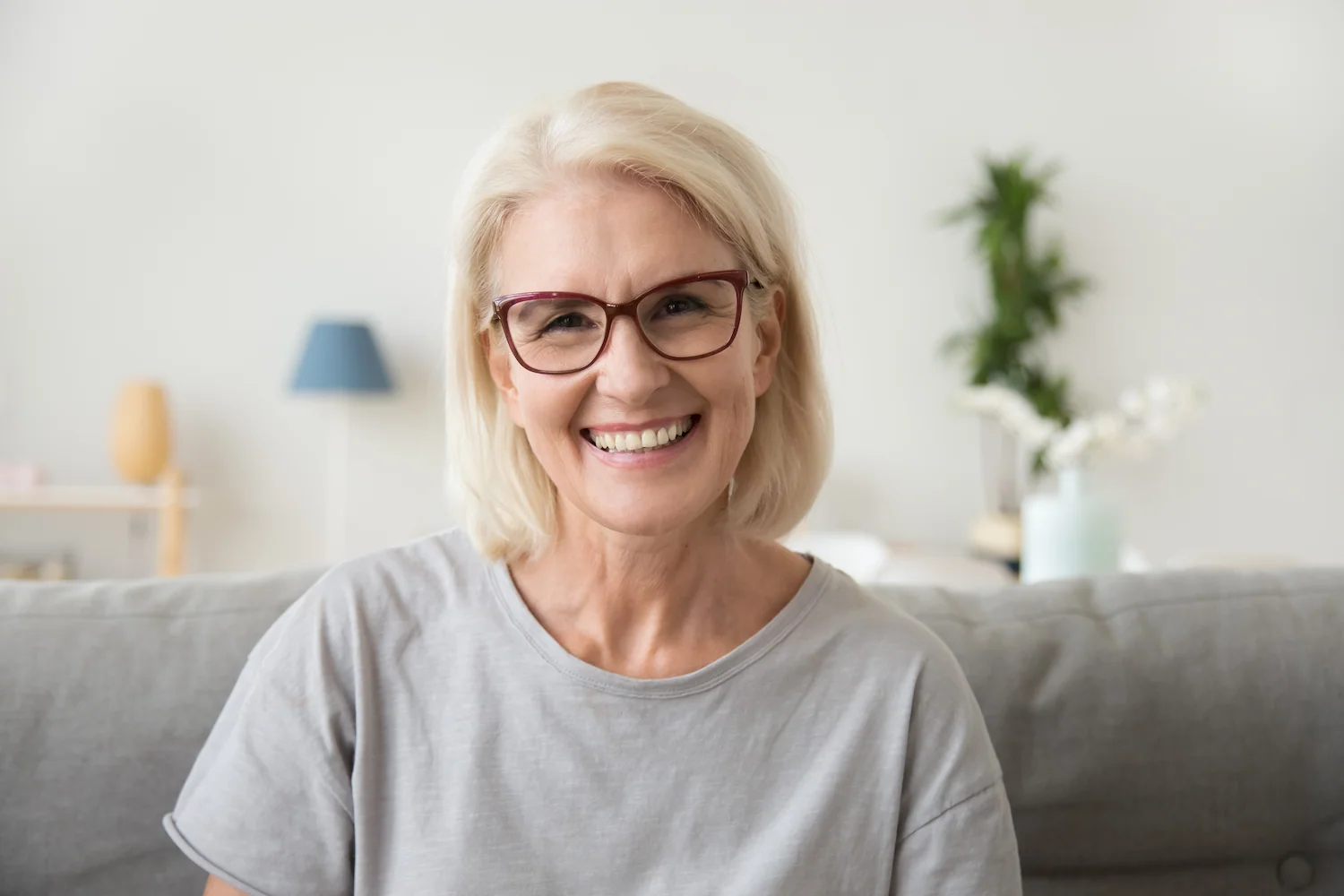Smiling older woman with short blonde hair and glasses, wearing a light gray shirt, sits on a gray sofa in a bright living room with plants and home decor in the background, embodying the spirit of wellness and anti-aging promoted by the Institute For Ageless Living.