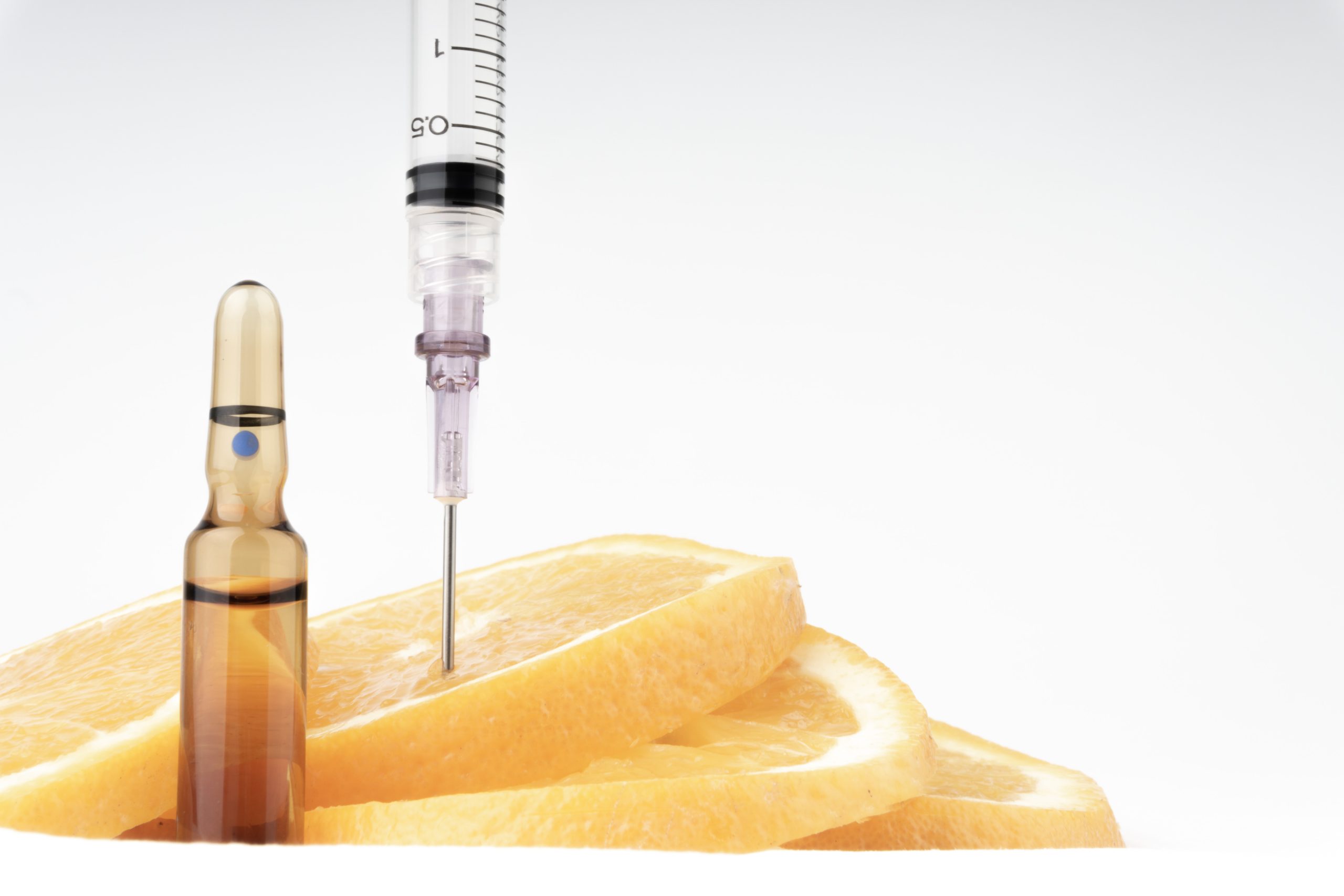 Several orange slices are stacked next to a glass ampoule, evoking a sense of wellness. A syringe needle is inserted into one of the orange slices, suggesting innovative approaches to anti-aging. The background is plain white, creating a clean and clinical aesthetic reminiscent of treatments at the Institute For Ageless Living.