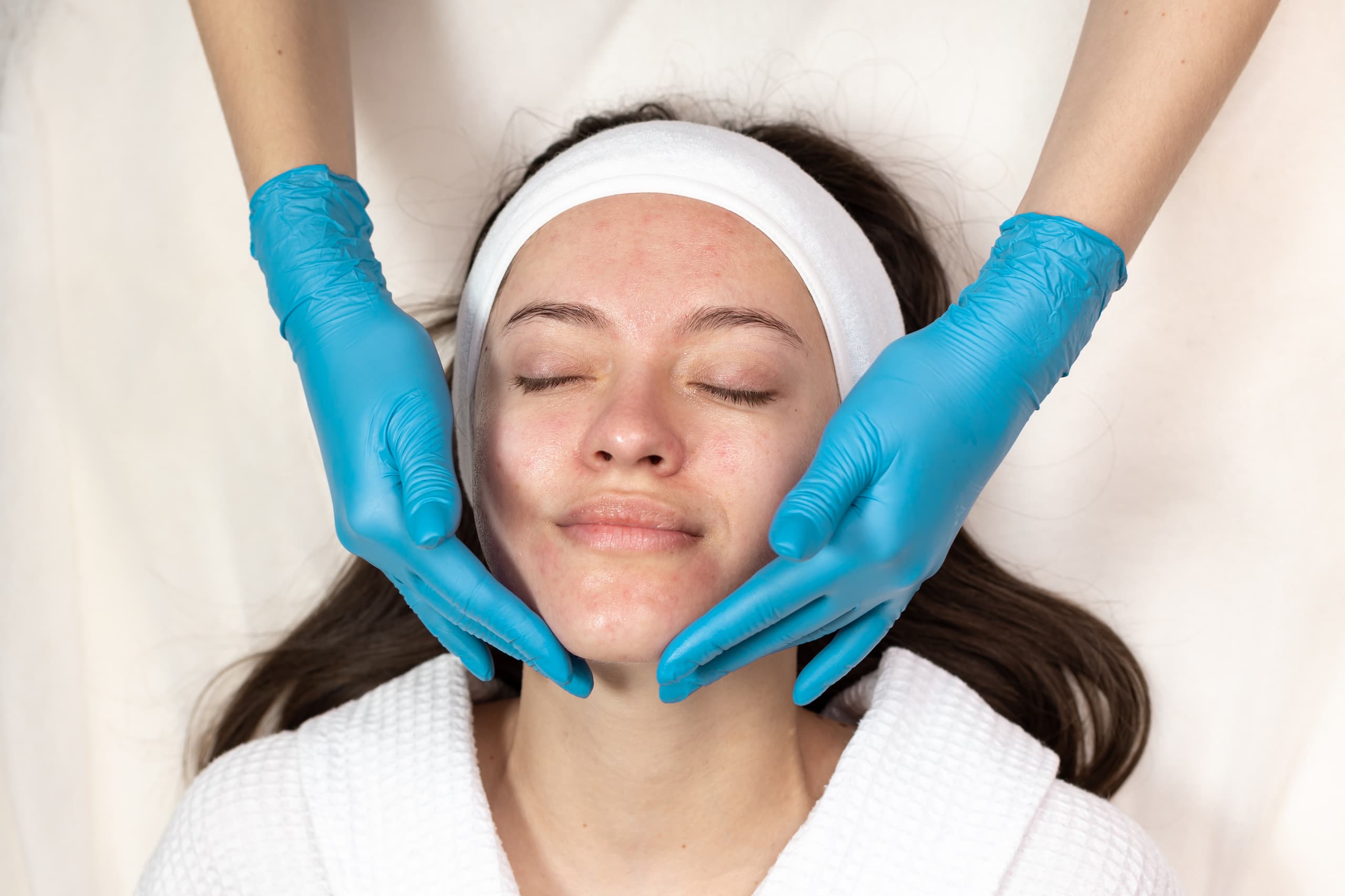 A person in a white robe with closed eyes receives an anti-aging facial treatment from someone wearing blue gloves. The person has a white headband and slightly red skin, reflecting the rejuvenating wellness services offered at the Institute For Ageless Living.