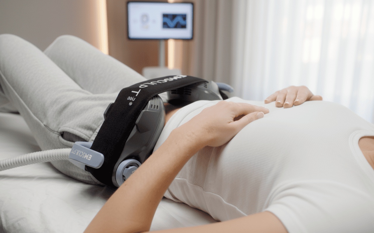 A person lies on a bed with a medical device belt strapped around their abdomen, likely receiving a wellness or body contouring treatment at the Institute For Ageless Living. A monitor and medical equipment are visible in the background.