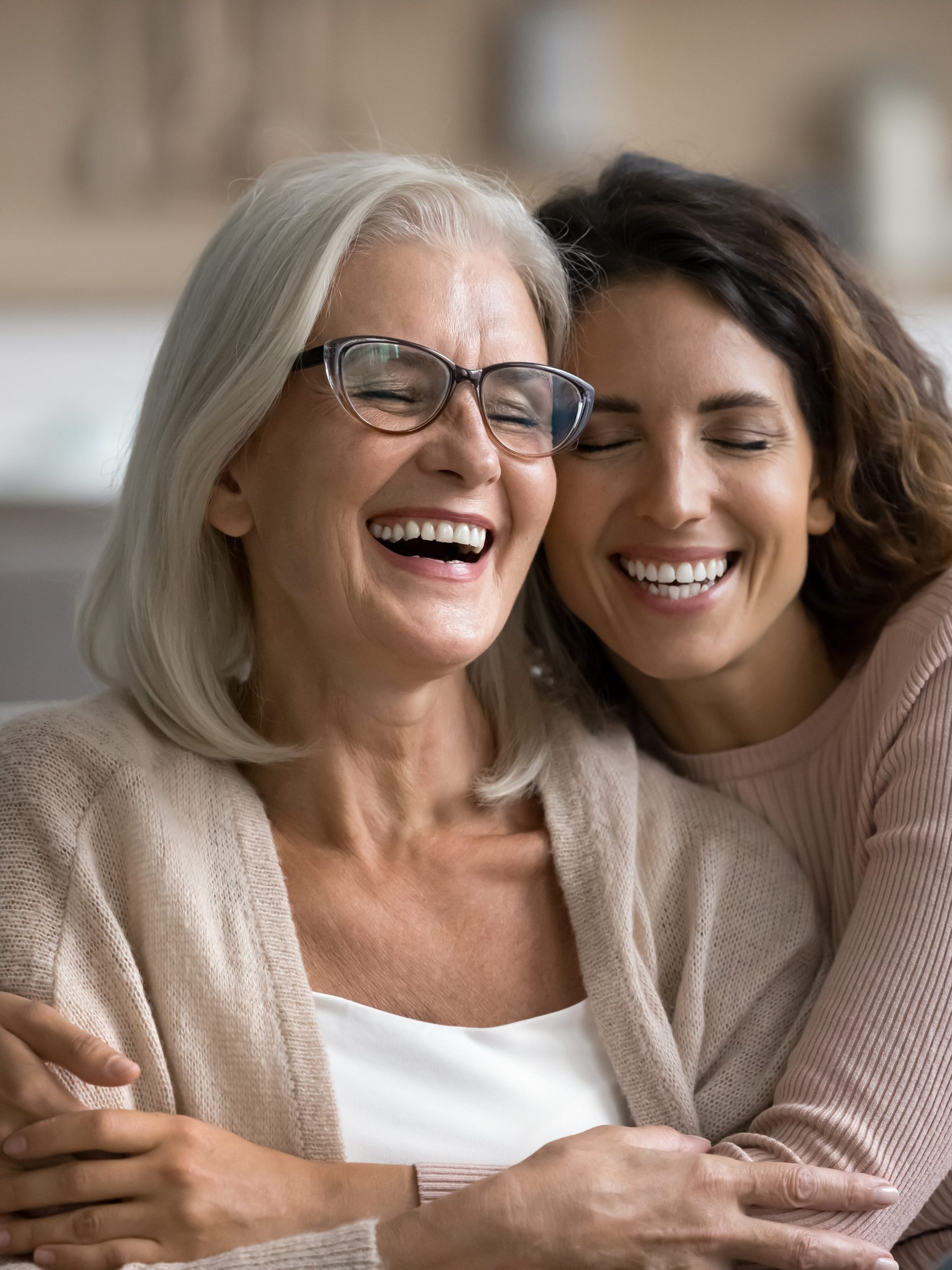 Two women, one older with gray hair and glasses, and one younger with brown hair, are smiling and hugging each other closely, expressing happiness and affection—a beautiful moment that reflects the spirit of wellness promoted by the Institute For Ageless Living.
