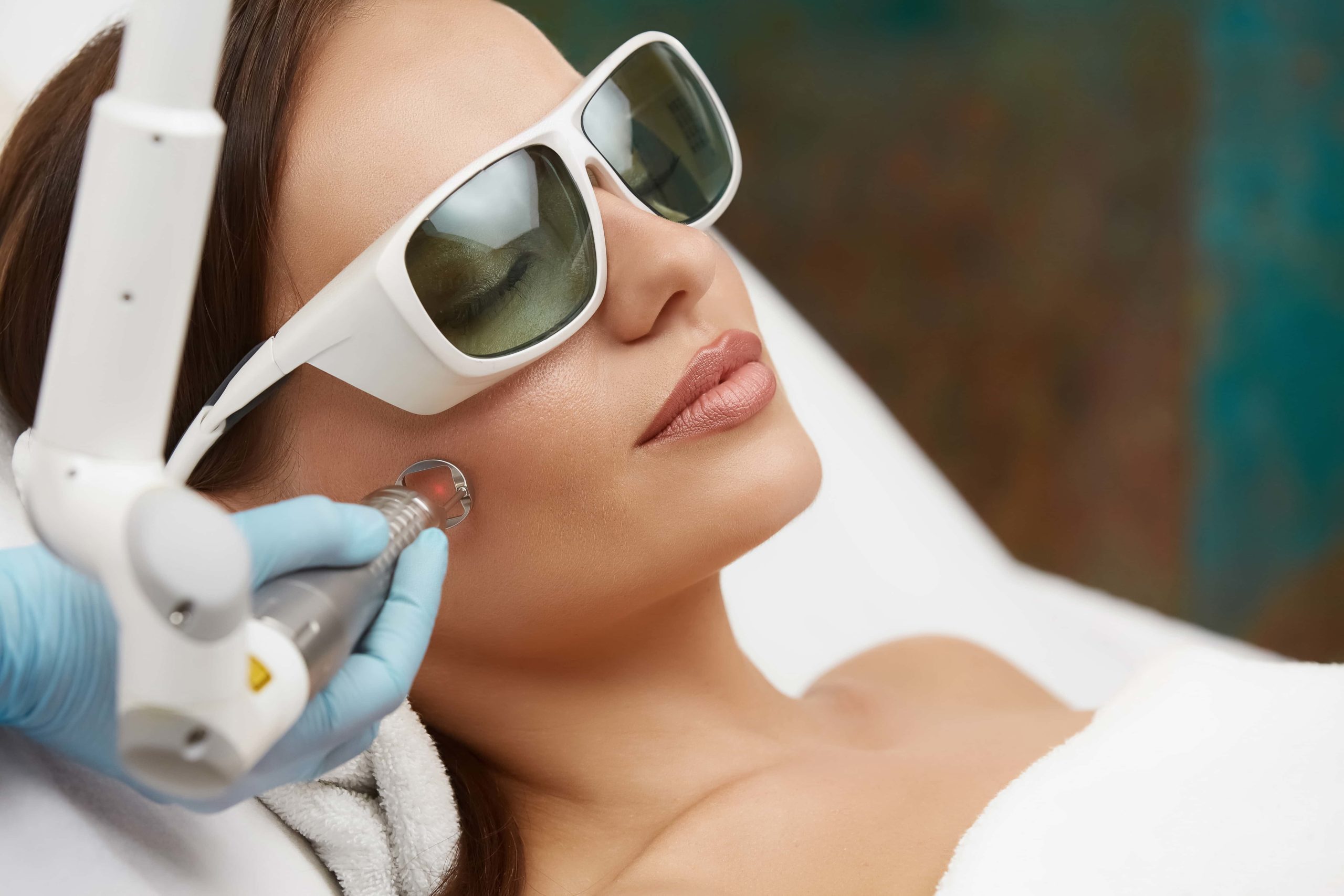 A woman wearing protective goggles receives a laser skin treatment on her face from a professional in blue gloves, highlighting the wellness expertise offered at the Institute For Ageless Living.