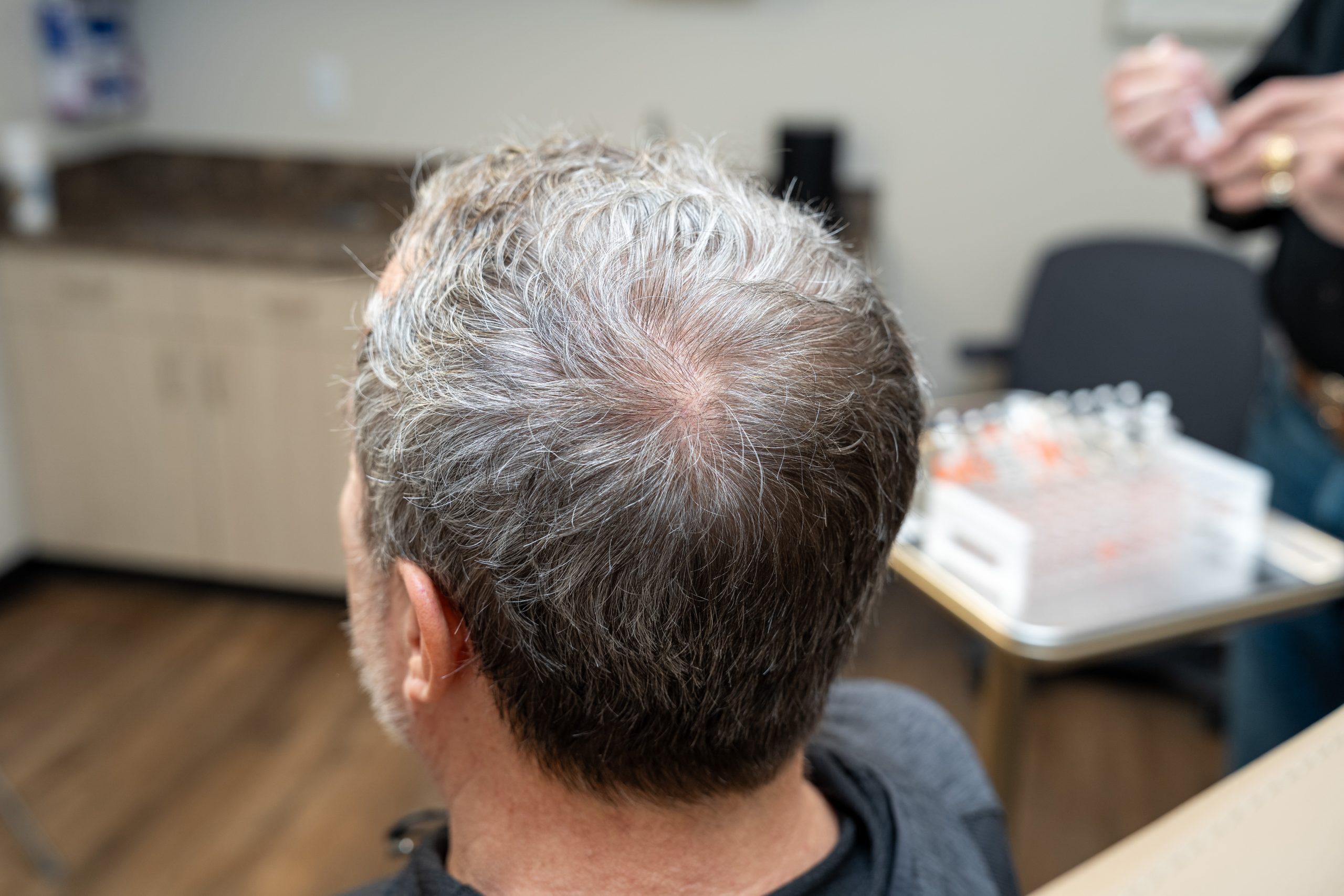 A man with thinning gray hair sits in a clinic, facing away. Nearby, a medical professional from the Institute For Ageless Living prepares an anti-aging injection. Wellness-focused medical supplies are arranged neatly on a table.
