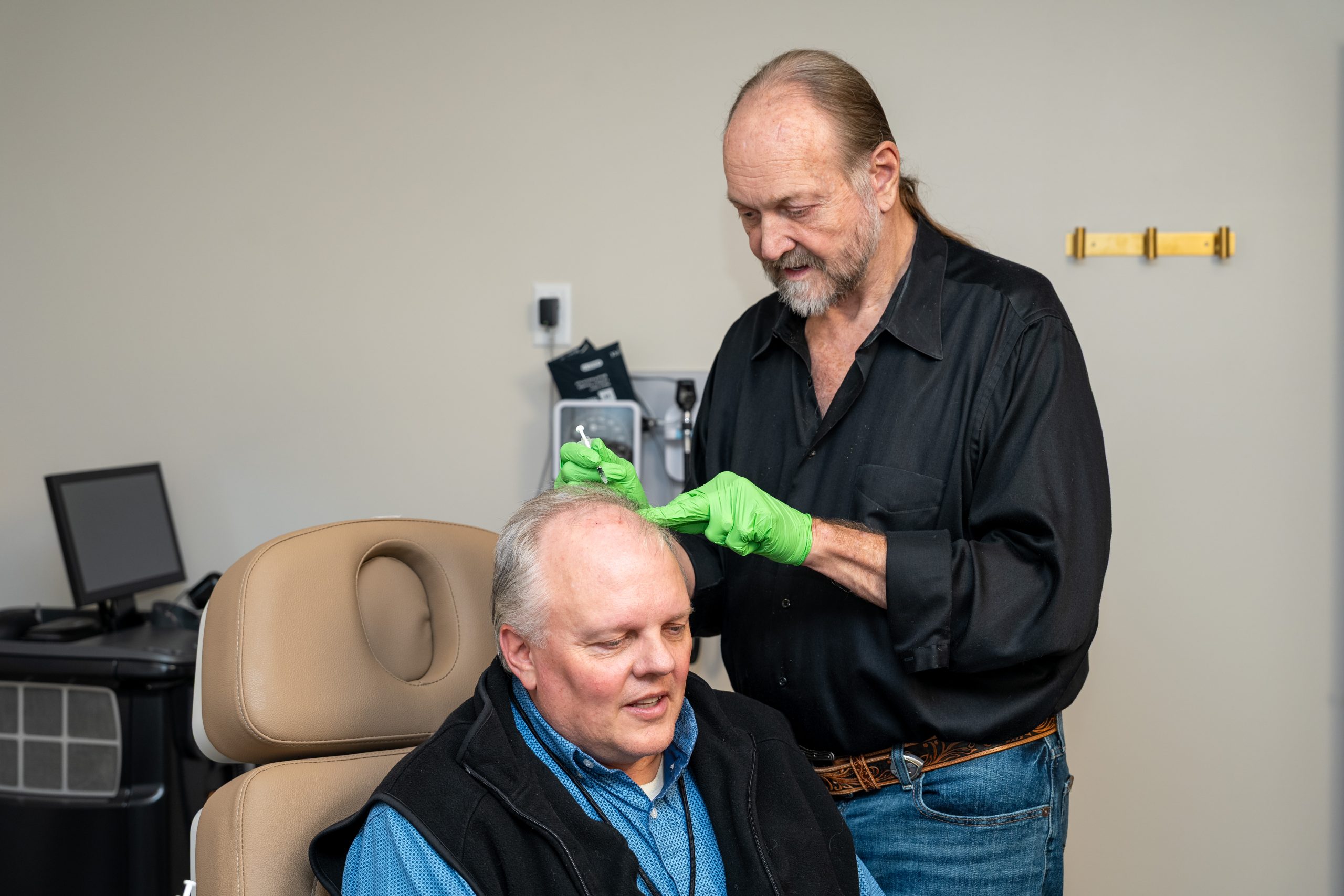 A man in a black shirt and green gloves examines or treats the scalp of a seated older man in a medical office setting, reflecting the Institute For Ageless Living's commitment to wellness and anti-aging care.