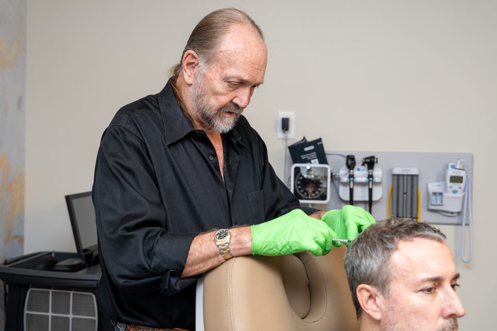 At the Institute For Ageless Living, a man wearing green gloves and a black shirt injects a needle into another man's scalp in a medical office setting, demonstrating their commitment to advanced wellness and anti-aging treatments.