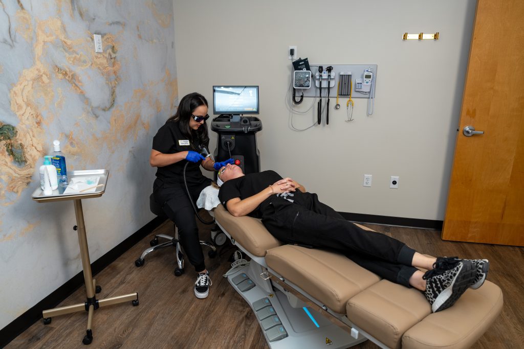 A medical professional in black scrubs uses equipment on a patient lying on a padded exam chair in a modern clinic room with medical tools and a computer nearby, exemplifying the Institute For Ageless Living's commitment to wellness and anti-aging care.