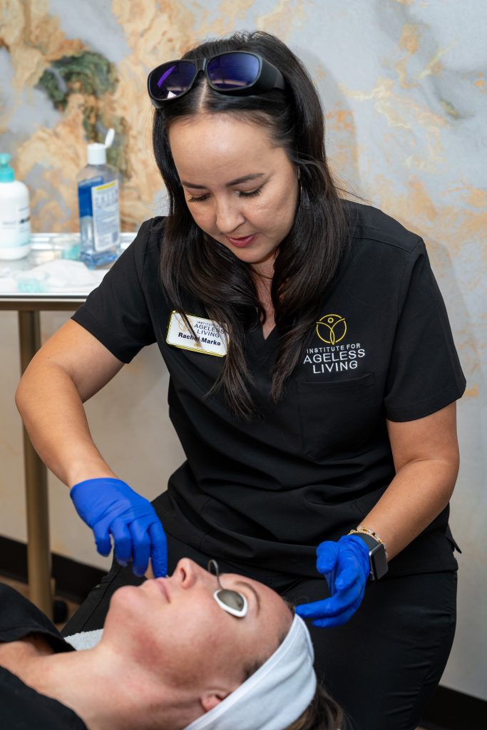 A woman in black scrubs and blue gloves uses a metal tool on a client's face during an anti-aging skincare treatment. The client lies on a table with a towel wrapped around their head, enjoying a moment of wellness. Skincare products are neatly arranged on a side table nearby at the Institute For Ageless Living.