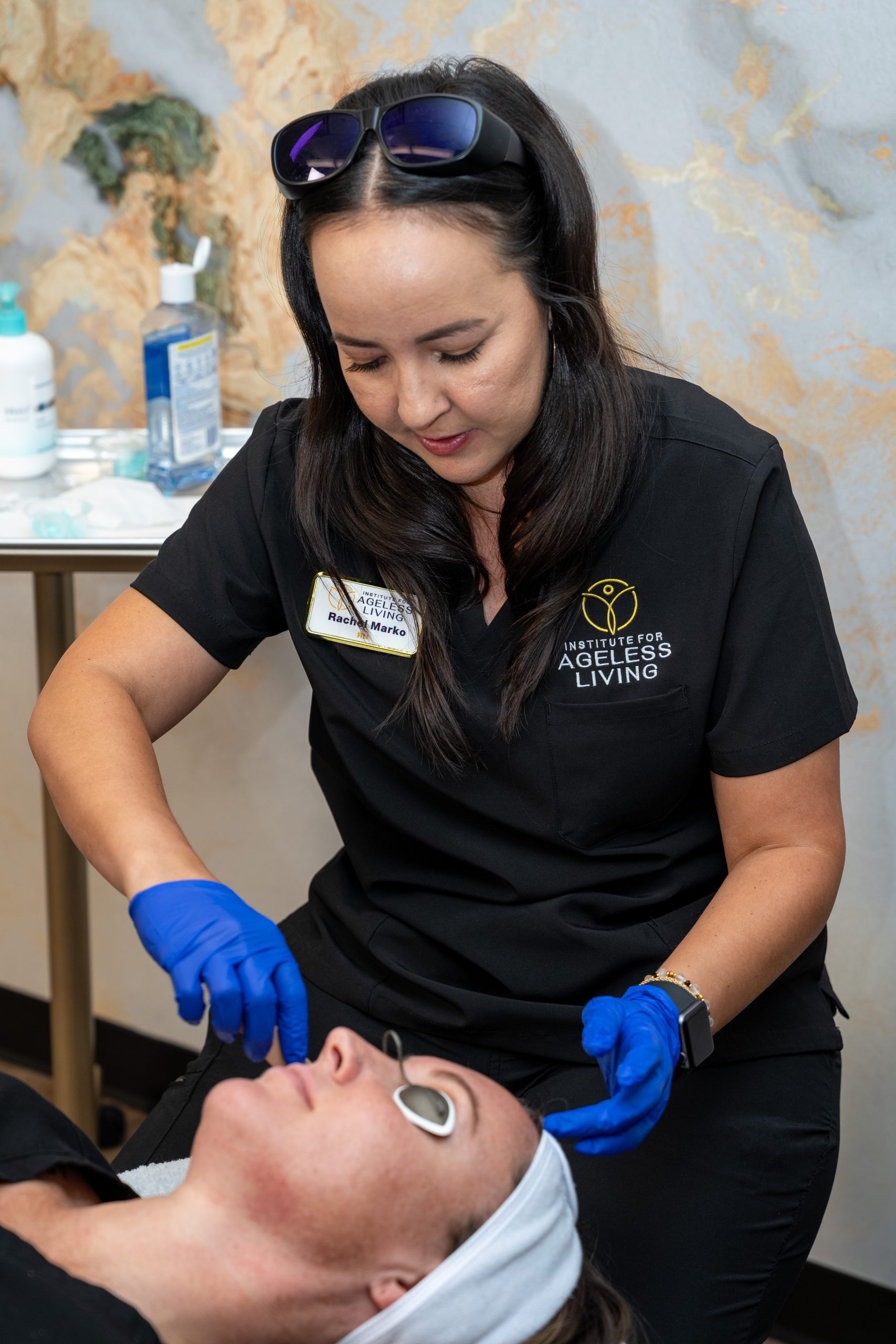 A woman in black scrubs and blue gloves uses a metal tool on a client's face during an anti-aging skincare treatment. The client lies on a table with a towel wrapped around their head, enjoying a moment of wellness. Skincare products are neatly arranged on a side table nearby at the Institute For Ageless Living.