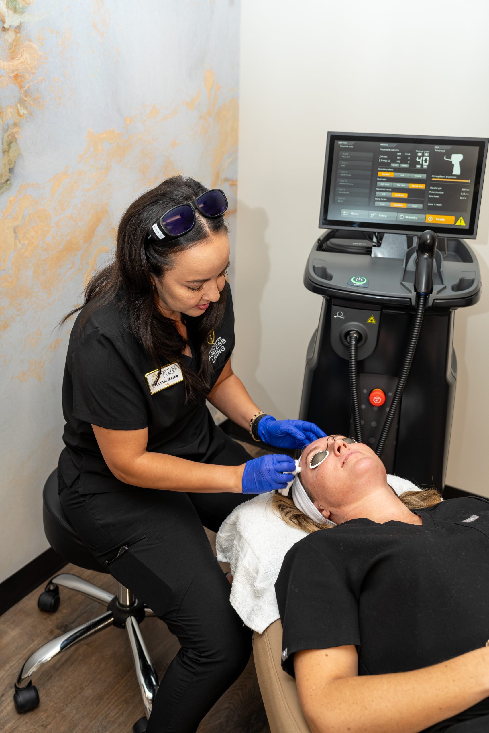A woman wearing gloves performs a facial laser treatment on a client lying down with eye protection, next to a laser machine displaying settings on its screen—an advanced anti-aging procedure at the Institute For Ageless Living, committed to promoting wellness and rejuvenation.