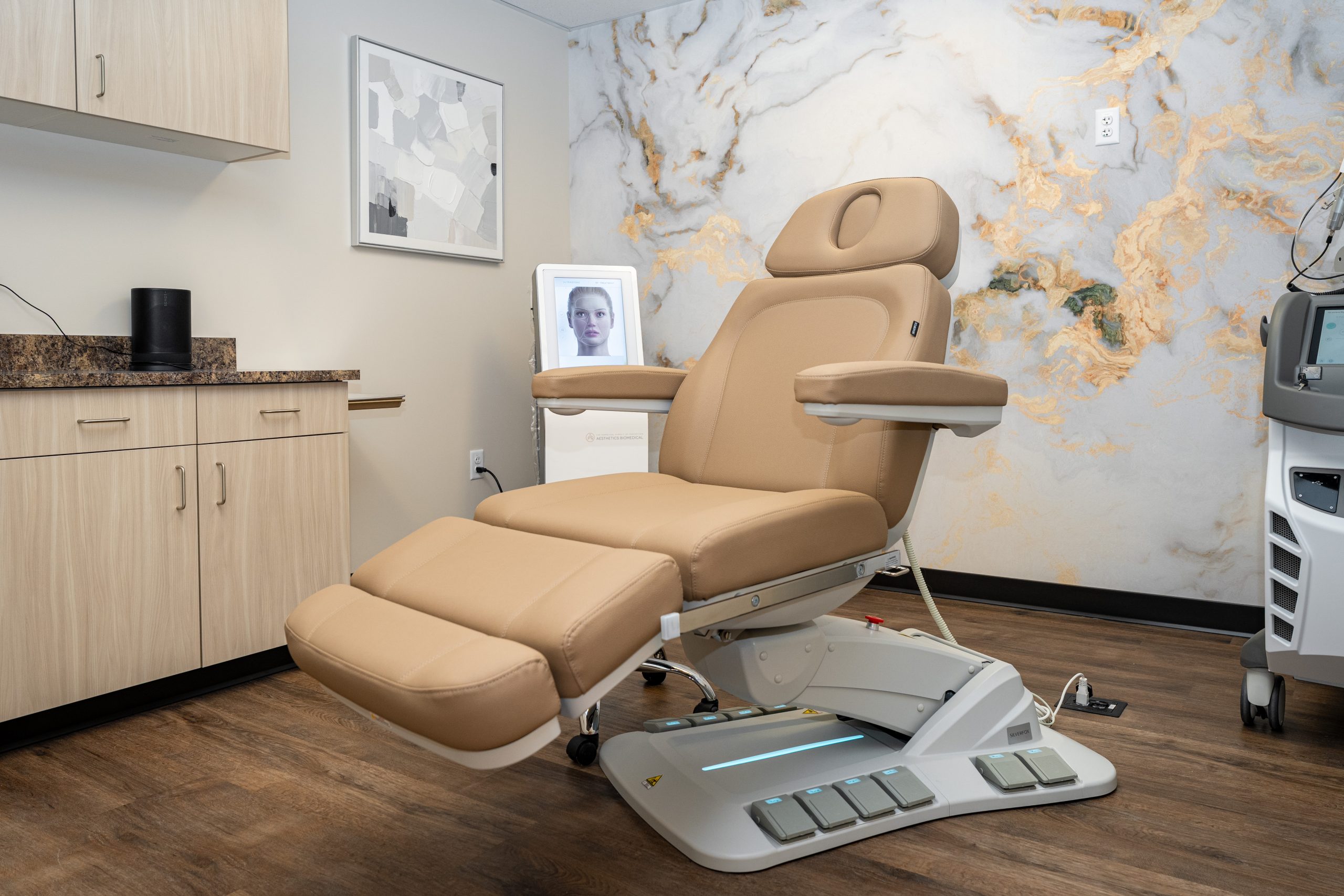 A modern medical exam chair in a clean clinic room with wood flooring, beige cabinetry, and marble-patterned walls creates an environment focused on wellness. In the background, a digital screen displays a woman's face, reflecting the Institute For Ageless Living's commitment to anti-aging care.