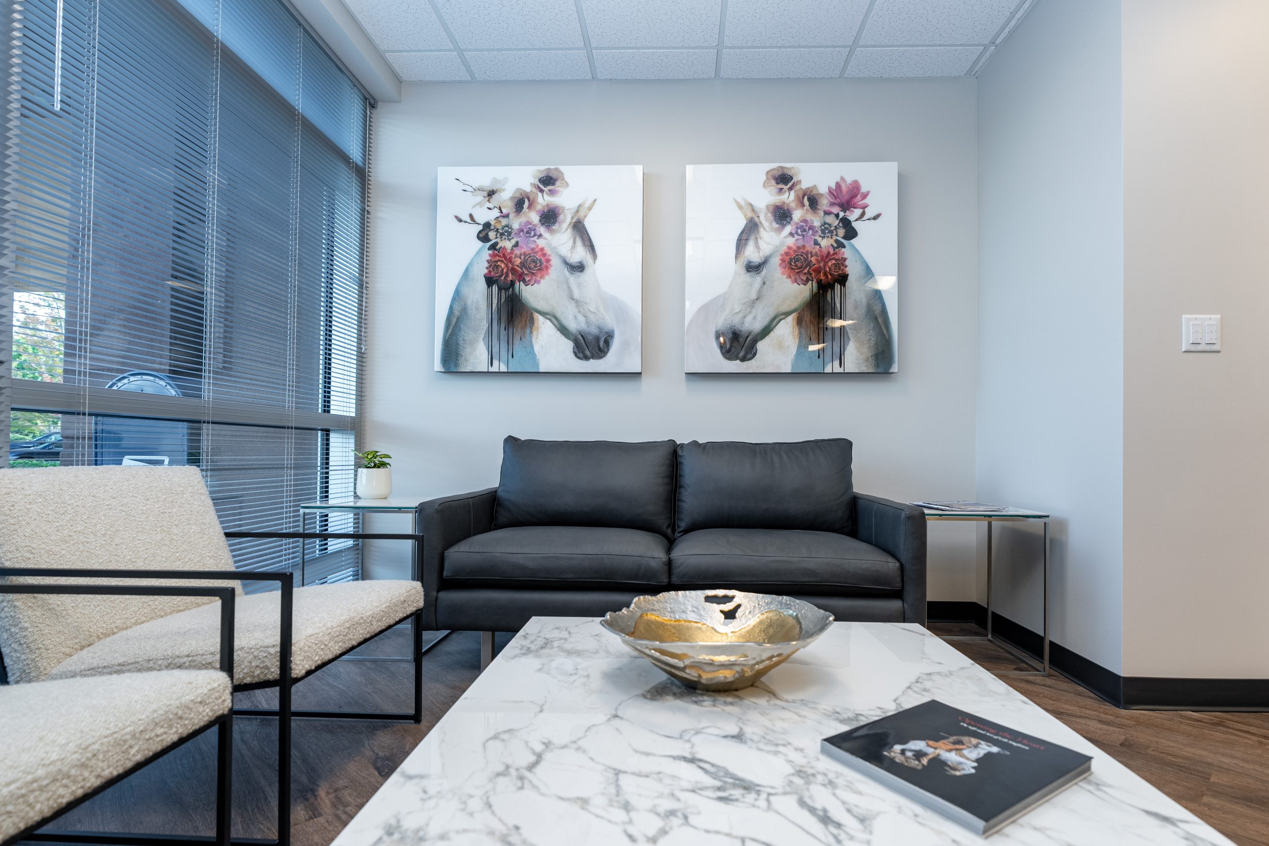 Modern waiting room with a black sofa, white armchair, marble coffee table, gold bowl, and two framed unicorn art pieces on the wall. Large windows let in natural light, creating an inviting atmosphere perfect for a wellness retreat at the Institute For Ageless Living.