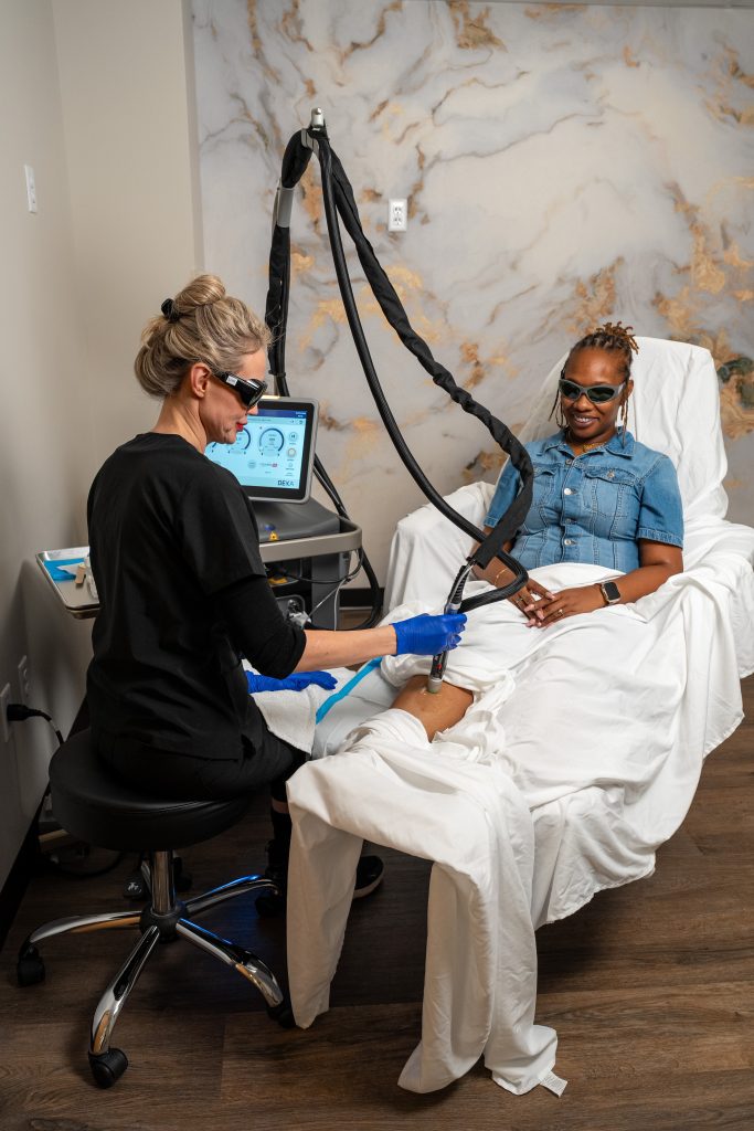 A woman in a denim shirt smiles while receiving a laser treatment on her leg from a technician wearing black scrubs and protective glasses in a medical office at the Institute For Ageless Living, where expert care is dedicated to wellness and anti-aging treatments.