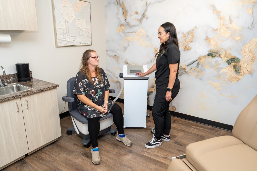 A woman in a floral top sits in a clinic chair while a healthcare worker in black scrubs stands by a medical device, both smiling in a modern medical office dedicated to anti-aging and wellness at the Institute For Ageless Living.