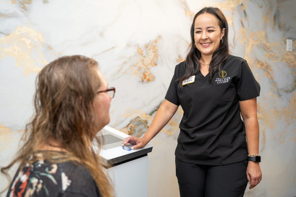 A smiling healthcare worker in black scrubs talks with a seated woman in a clinic setting, embodying the spirit of wellness promoted by the Institute For Ageless Living. The wall behind them is light with a marbled pattern.