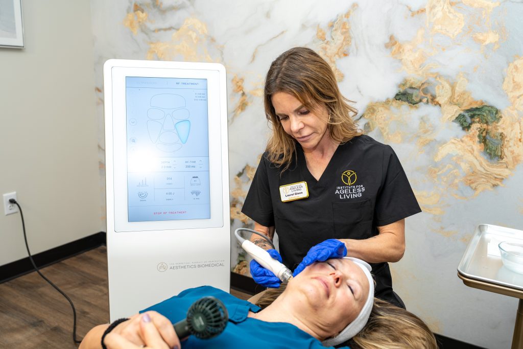 A woman in scrubs uses a medical device on a patient's forehead in a clinic room at the Institute For Ageless Living. A screen next to them displays facial diagrams, reinforcing the clinic's focus on anti-aging and overall wellness. The patient lies on a treatment table, holding a small blue tool.