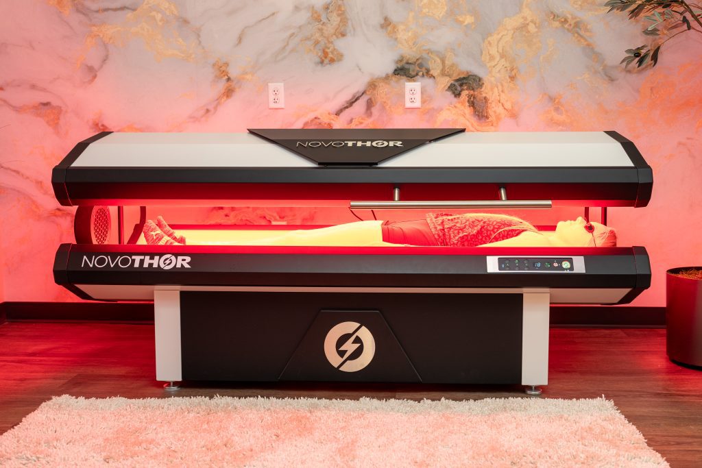 A person lies inside a Novothor red light therapy bed in a spa-like room with marble walls and a white rug, bathed in red light—a serene scene at the Institute For Ageless Living, where innovative wellness and anti-aging therapies are seamlessly woven into every experience.