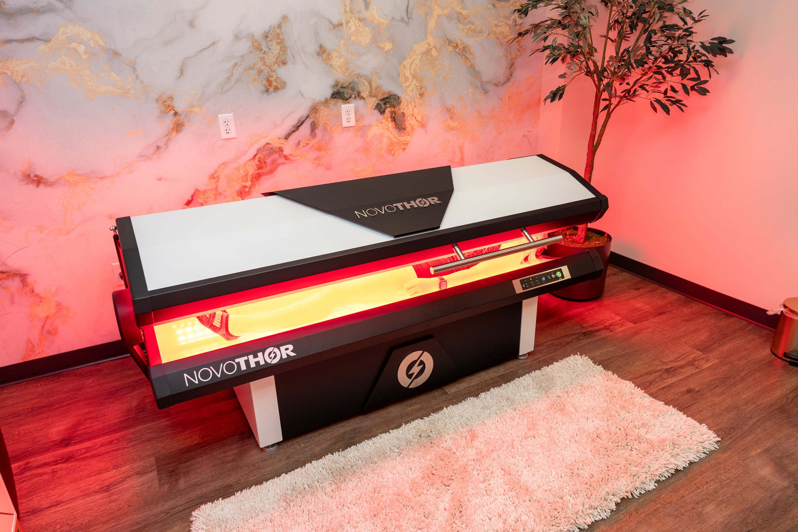 A NovoTHOR light therapy bed with red interior lights is positioned on a wooden floor beside a white rug and a potted plant, against a marble-patterned wall. This wellness device, offered by the Institute For Ageless Living, creates an inviting space designed to support anti-aging benefits and overall vitality.
